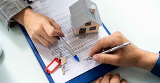 A man signing a piece of paper. A set of keys and a miniature house rest on top of the paper